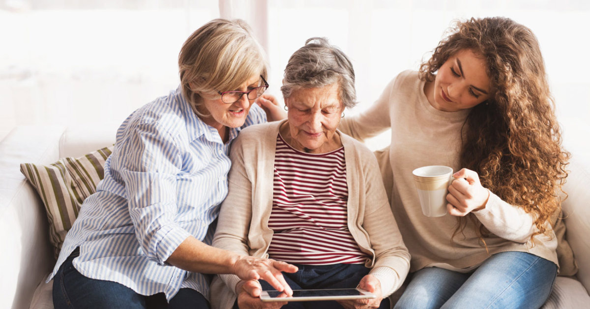Elderly woman with daughter and granddaughter reading aged care news on tablet while enjoying coffee together